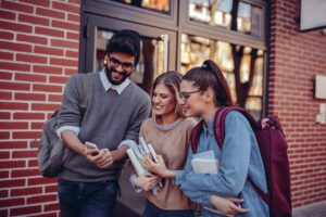 Three,Students,Walking,Down,The,Street,Looking,At,A,Mobile