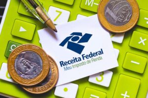 Logotipo da Receita Federal sobre uma calculadora verde ao lado de moedas de real, representando declaração de Imposto de Renda.