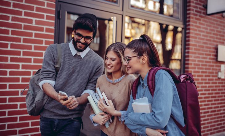 Three,Students,Walking,Down,The,Street,Looking,At,A,Mobile